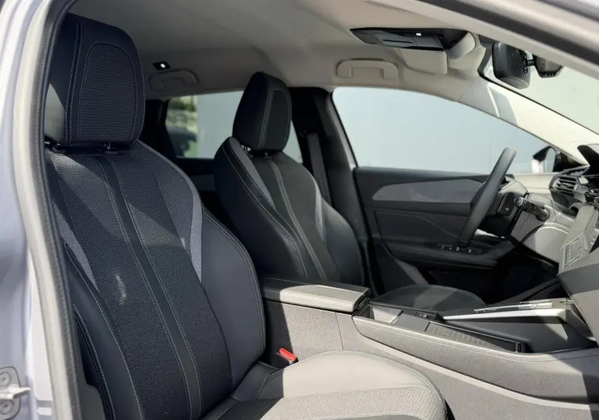 Vue intérieure côté conducteur de la Peugeot 308 SW 2025, sièges tri matières gris avec surpiqûres bleues et accoudoir central.