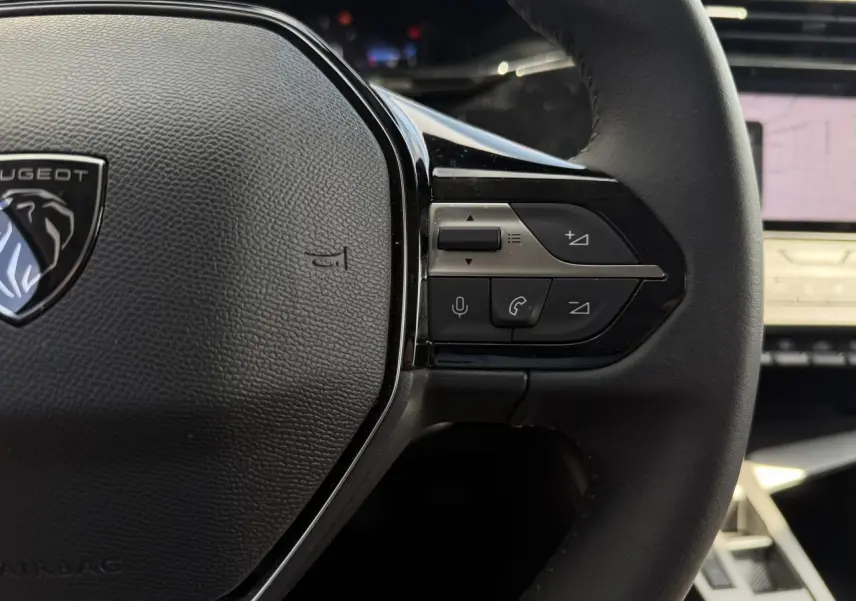 Gros plan sur le volant cuir noir du Peugeot 308 SW 2025 avec commandes intégrées et écran tactile flou en arrière-plan.