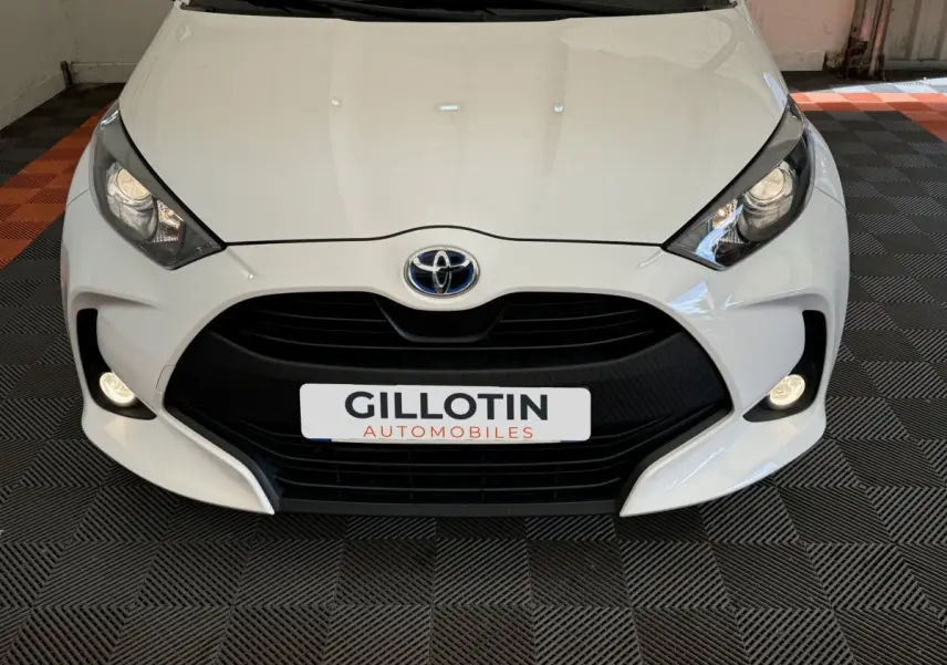 Vue frontale rapprochée d'une Toyota Yaris hybride blanche avec calandre noire et phares allumés.