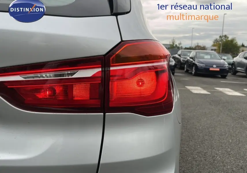 Gros plan sur le feu arrière droit d’un BMW X1 gris argent métal, avec un parking et plusieurs voitures en arrière-plan.
