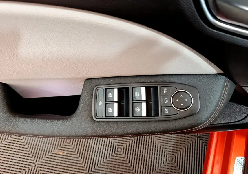 Gros plan sur la commande des vitres côté conducteur avec surpiqûres orange, intérieur clair et carrosserie orange visible.
