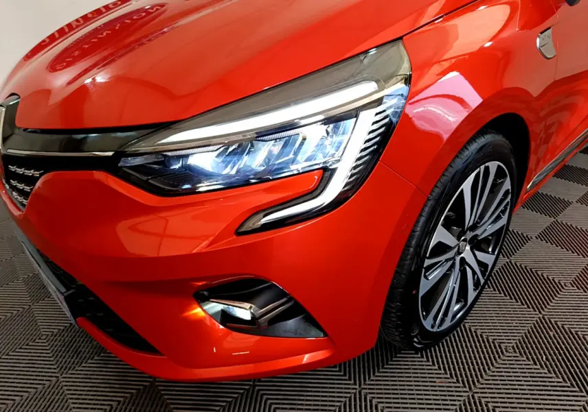 Gros plan sur l'avant droit orange vif de la Renault Clio Initiale Paris 2021, mettant en valeur le phare LED et la jante stylée.