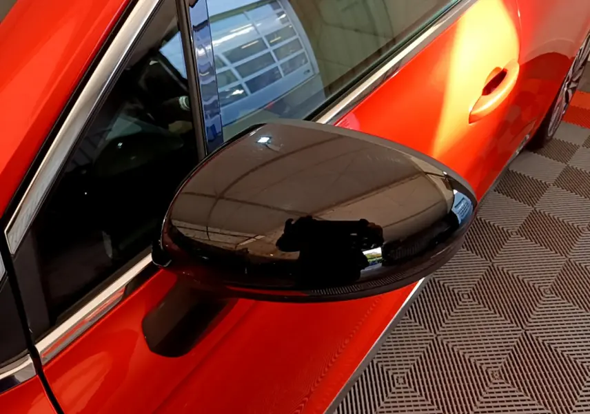 Rétroviseur noir brillant côté gauche d'une Renault Clio orange vue en gros plan sur fond de sol à motifs.