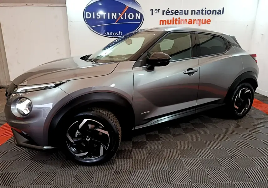 Nissan Juke Hybrid gris foncé vue de profil côté gauche, avec jantes noires et logo Distinxion en arrière-plan.