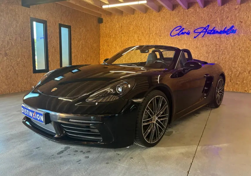 Porsche 718 Boxster 2.0 300 noir vue 3/4 avant droit en intérieur avec jantes alliage et capote ouverte.