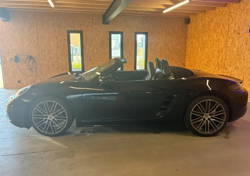 Vue de profil côté gauche d'une Porsche 718 Boxster noire cabriolet, avec jantes alliage et intérieur apparent.