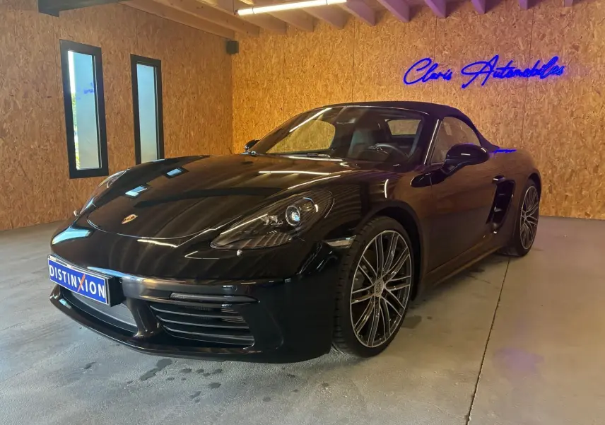 Porsche 718 Boxster noir vue 3/4 avant droit en intérieur, avec jantes alliage et capote fermée.