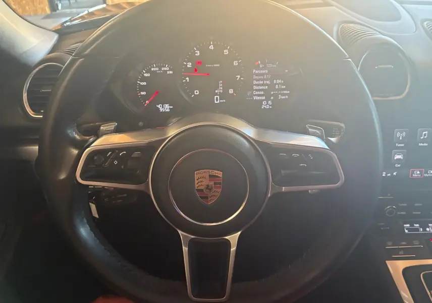 Vue rapprochée du volant noir de la Porsche 718 Boxster 2.0 300 de 2018 avec tableau de bord numérique allumé.
