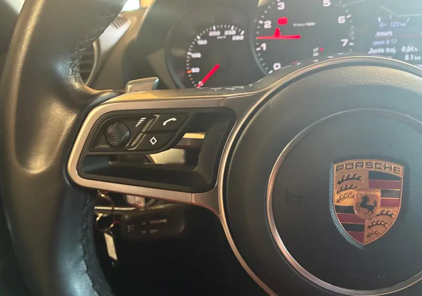 Gros plan sur le volant noir d’un Porsche 718 Boxster 2.0 300 de 2018 avec commandes intégrées et tableau de bord lumineux.