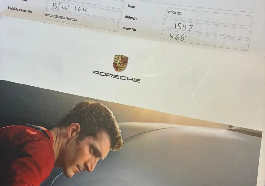 Homme en train de polir le capot avant noir d'une Porsche 718 Boxster 2.0 300 vue de face, mettant en valeur l'écusson Porsche.