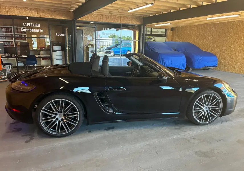 Vue de profil côté gauche d'une Porsche 718 Boxster noire cabriolet, avec jantes alliage et intérieur visible.