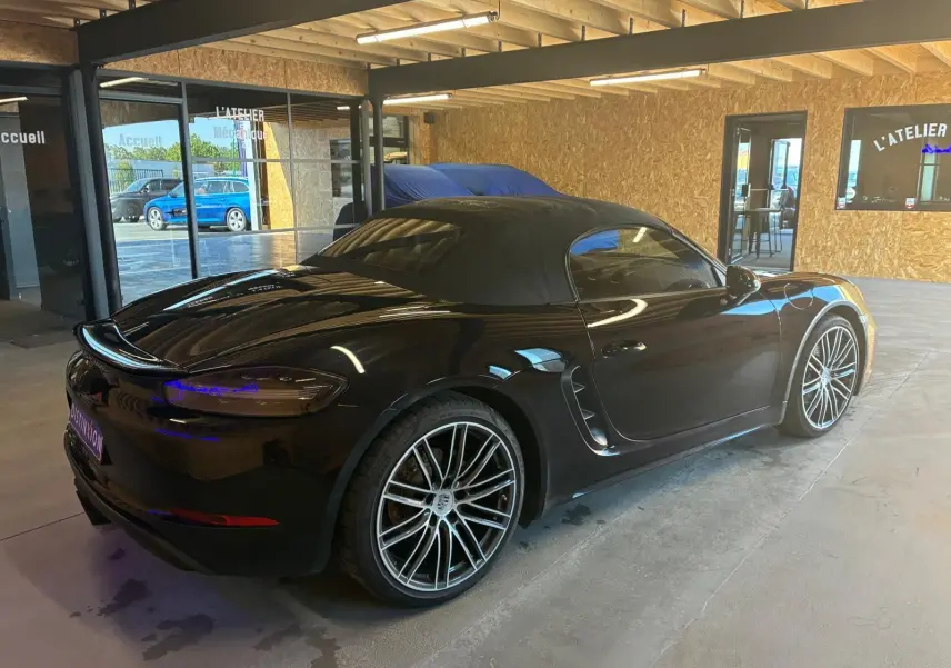 Porsche 718 Boxster 2.0 300 noir vue 3/4 arrière droit en intérieur avec toit souple fermé et jantes multibranches.