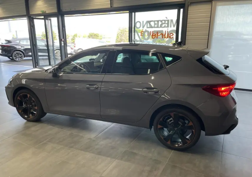 Vue de profil côté gauche d'une CUPRA Leon gris Graphène avec jantes alliage 18'' cuivrées en intérieur showroom.