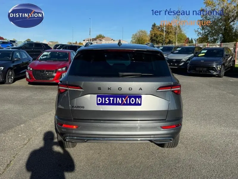 Vue arrière d'un SKODA KAROQ gris perle avec feux LED et barres de toit visibles sur un parking extérieur.