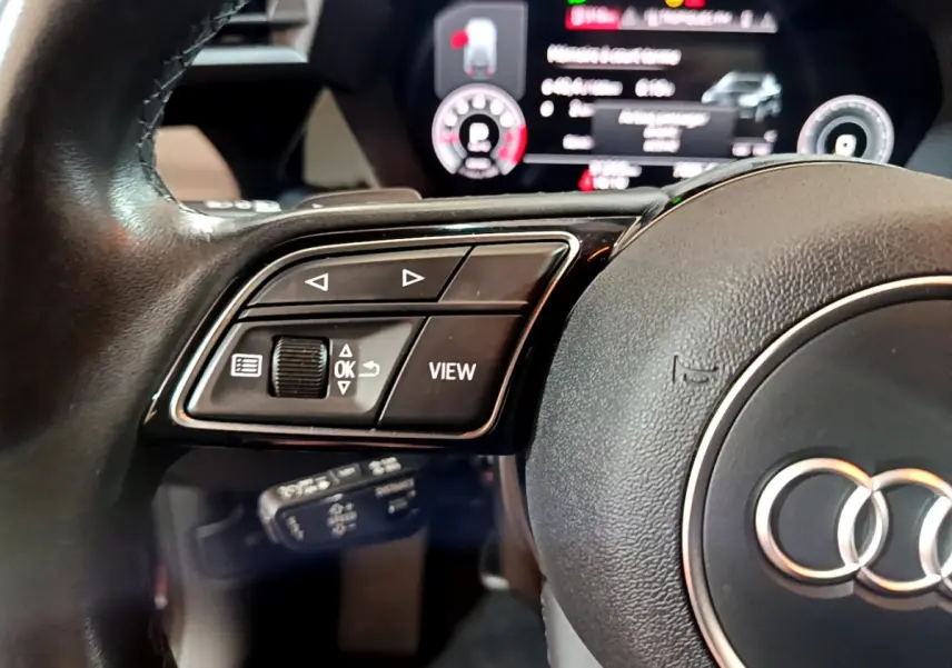 Gros plan sur la partie gauche du volant cuir d'une Audi A3 35 TFSI 2023 avec commandes multifonctions et tableau de bord numérique flou.