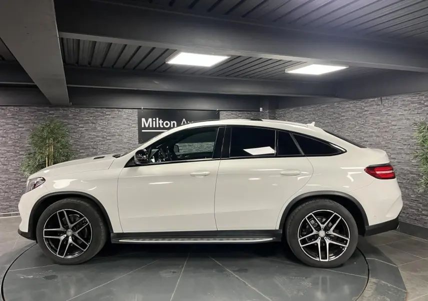 Profil côté gauche du Mercedes GLE Coupé 350 d blanc avec jantes AMG noires et toit panoramique en intérieur showroom.