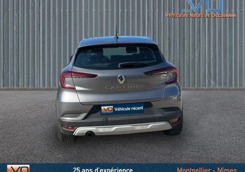 Vue arrière d'un Renault Captur gris 2021 avec feux allumés et plaque "Véhicule récent" sur fond industriel.