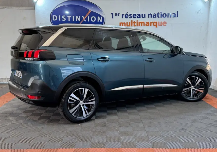 Vue latérale droite d’un Peugeot 5008 bleu métallisé avec toit noir et jantes alliage bicolores, en intérieur showroom.