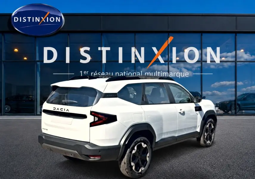 Vue 3/4 arrière droite du SUV blanc Dacia Bigster 2026 avec barres de toit noires et jantes alu distinctives.