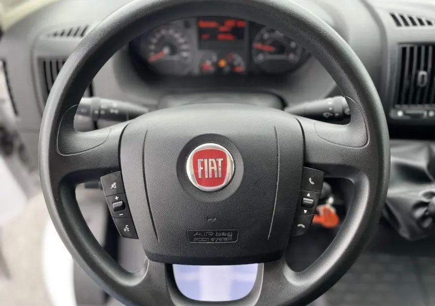 Volant noir avec logo FIAT rouge au centre, tableau de bord flou en arrière-plan dans un Fiat Ducato Fourgon blanc.