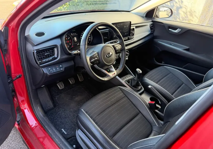 Intérieur côté conducteur d'une KIA RIO rouge grenadine 2022, montrant tableau de bord, volant multifonction et sièges noirs.