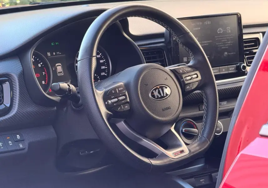 Vue intérieure centrée sur le volant cuir noir de la Kia Rio rouge grenadine, avec tableau de bord et écran tactile visibles.