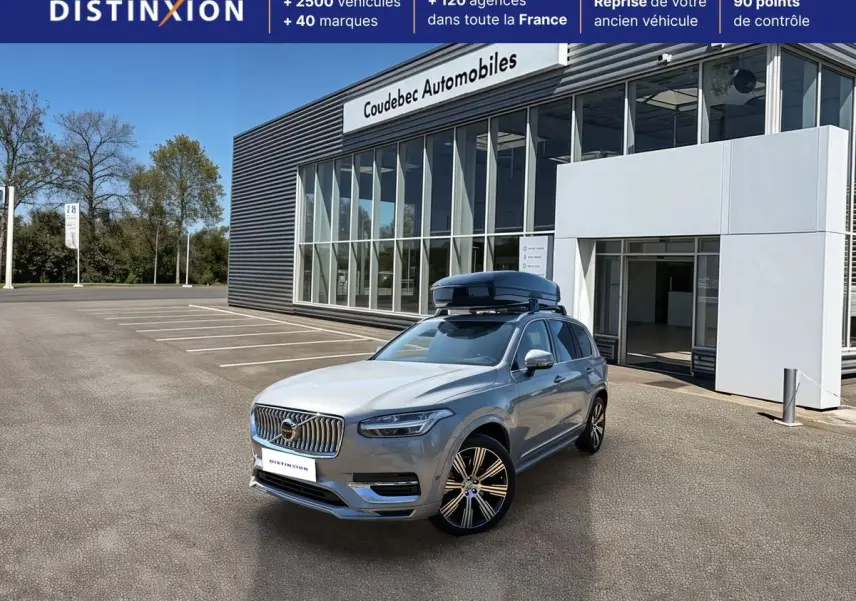 Vue 3/4 avant droite du Volvo XC90 gris avec coffre de toit noir sur parking devant concession.