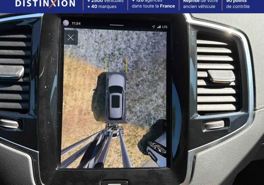 Vue intérieure de l'écran central montrant la caméra surround view 360° d'un Volvo XC90 gris stationné sur un sol mixte.