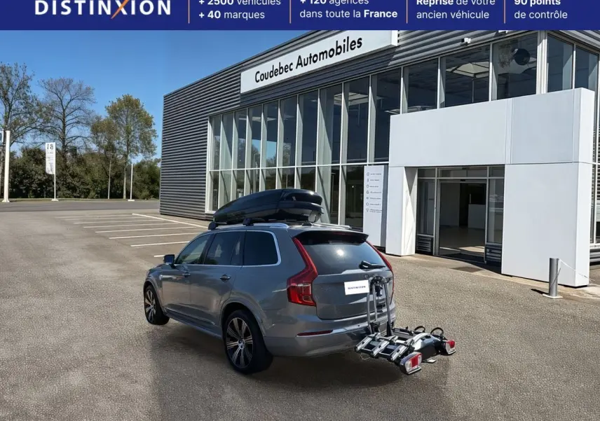 Vue 3/4 arrière d'un Volvo XC90 gris avec coffre de toit noir et porte-vélos fixé à l'attelage, devant un bâtiment commercial.