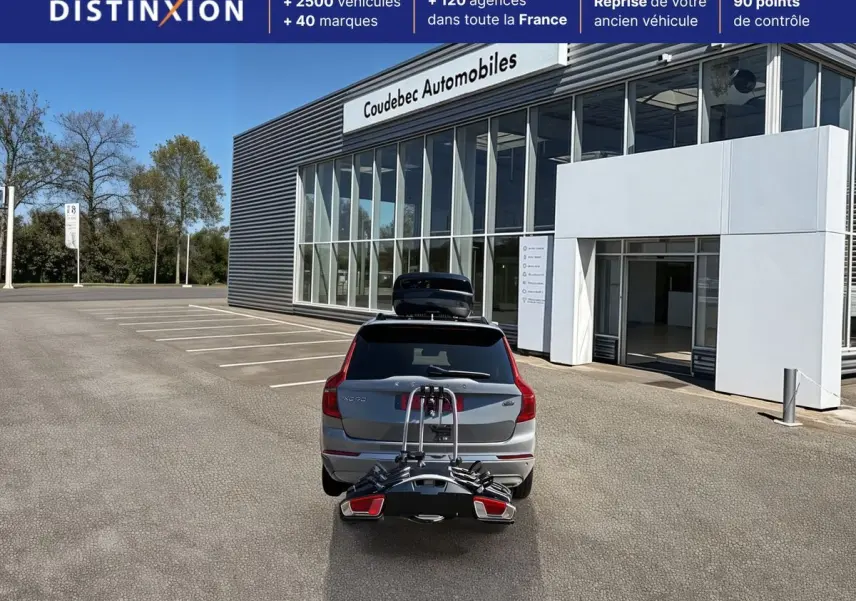 Vue arrière d'un Volvo XC90 gris avec coffre de toit et porte-vélos, stationné devant un bâtiment commercial.