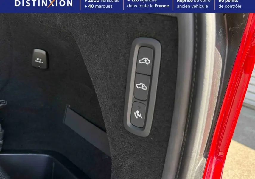 Gros plan sur les commandes intérieures du coffre d'un Volvo XC90 gris, avec bouton rabattage sièges et prise 12V visible.