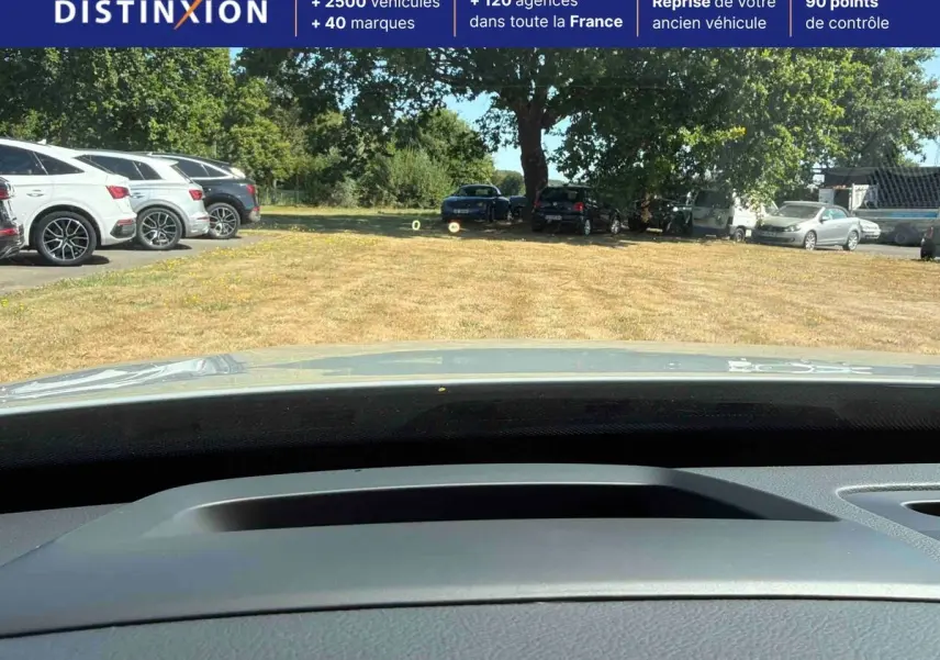 Vue depuis l'intérieur du Volvo XC90 gris 2024, montrant le tableau de bord et le pare-brise sur un parking extérieur.