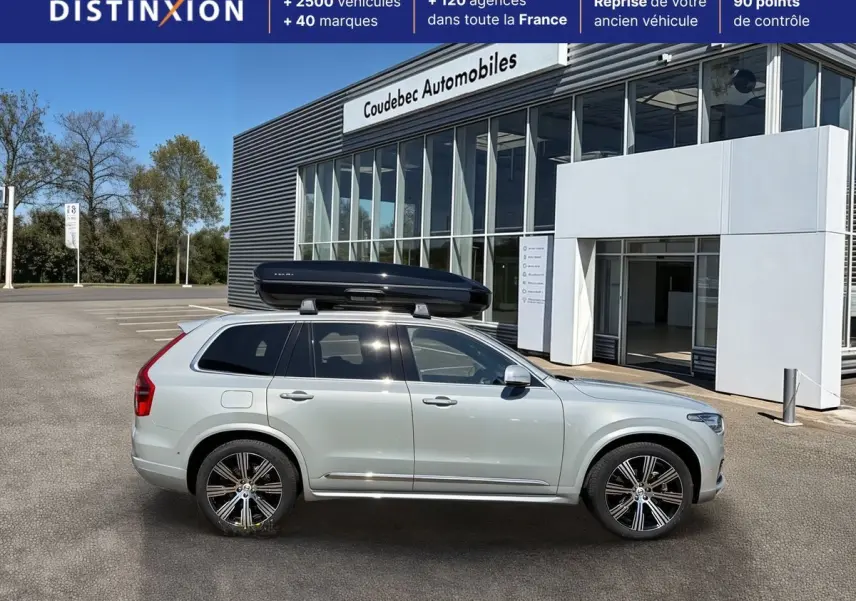 Vue de profil droit du Volvo XC90 gris clair avec coffre de toit noir sur un parking devant un bâtiment commercial.