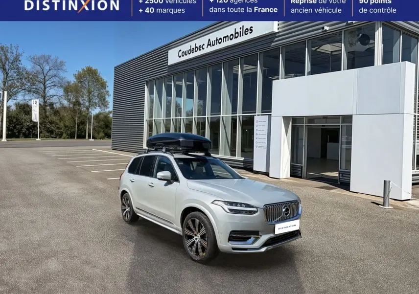 Vue 3/4 avant droite du Volvo XC90 gris avec coffre de toit noir, stationné devant un bâtiment commercial.