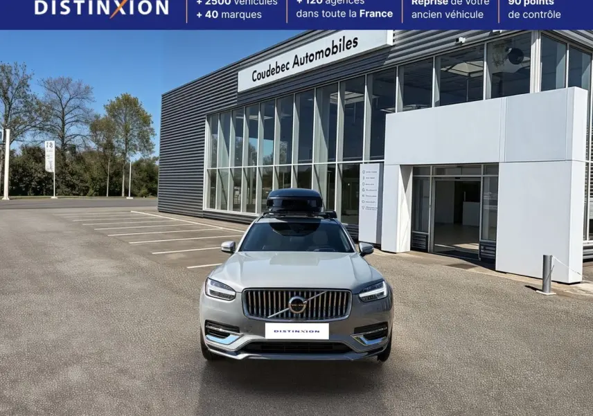 Vue avant d'un Volvo XC90 gris avec coffre de toit noir, stationné devant un bâtiment commercial.