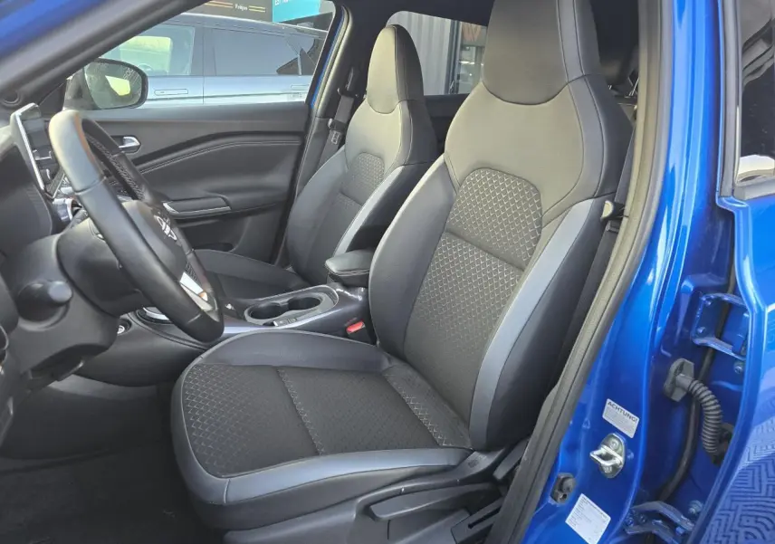 Vue intérieure côté conducteur du Nissan Juke II bleu magnetic, montrant les sièges avant en TEP noir et gris avec accoudoir central.