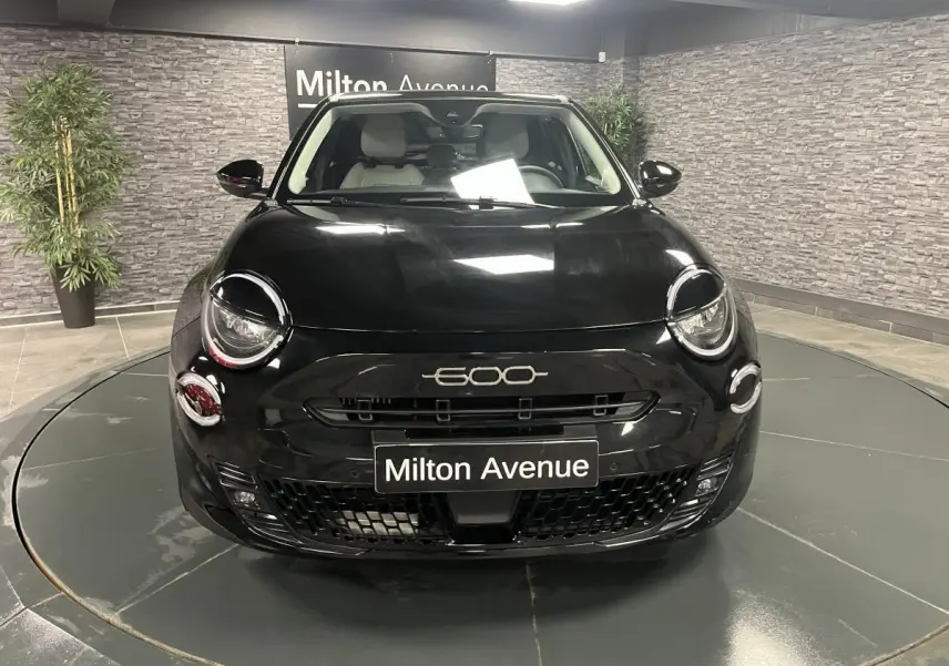 Vue frontale d'une FIAT 600 noire 2024 avec logo 600 chromé et plaque Milton Avenue en intérieur showroom.