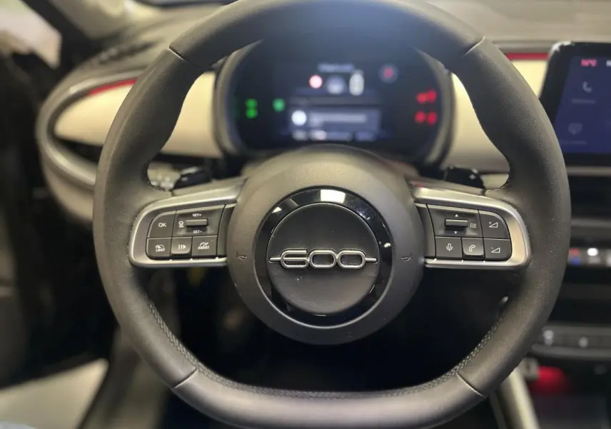 Gros plan sur le volant noir du Fiat 600 La Prima 2024 avec logo 600 et commandes intégrées, planche de bord ivoire en arrière-plan.