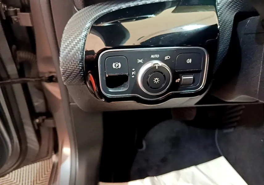 Gros plan sur le panneau de commande des phares et frein à main électrique côté gauche dans une Mercedes Classe A gris foncé.