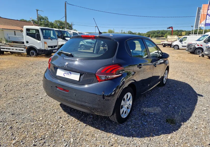 Peugeot 208 gris Hurricane vue en 3/4 arrière droit sur un terrain gravillonné avec plusieurs véhicules utilitaires en arrière-plan.