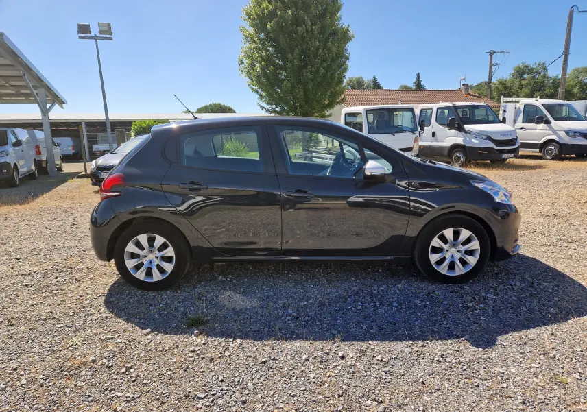 Peugeot 208 gris hurricane vue de profil côté gauche sur un sol gravillonné avec plusieurs véhicules utilitaires en arrière-plan.