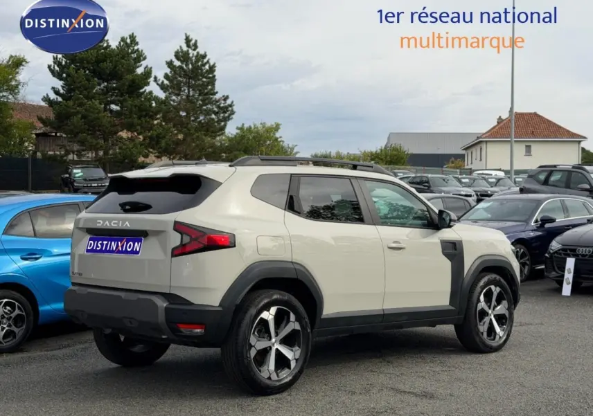 Vue 3/4 arrière droite d'un Dacia Duster 2025 Sandstone avec jantes alu et barres de toit visibles en extérieur.
