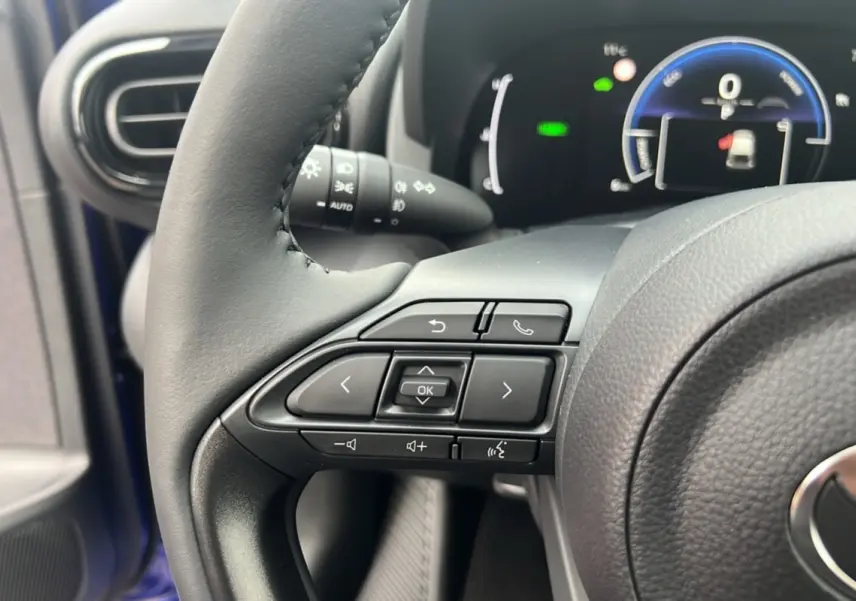 Gros plan sur les commandes au volant gauche de la Toyota Yaris 2025 bleu genievre, avec tableau de bord numérique en arrière-plan.