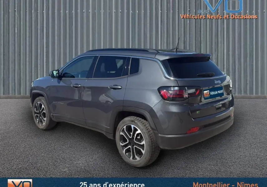 Jeep Compass gris 3/4 arrière droit avec barres de toit et jantes 18 pouces, stationné devant un mur métallique.