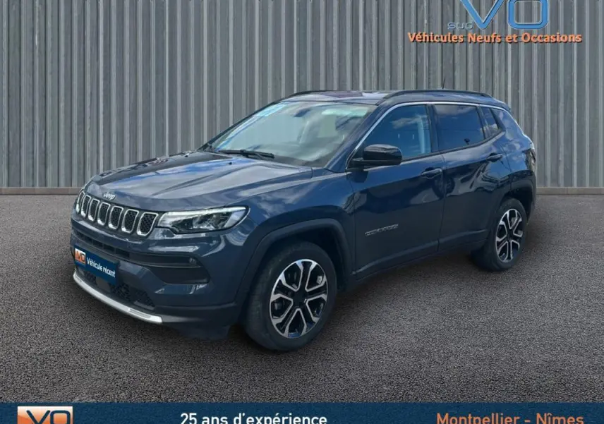 Jeep Compass bleu en 3/4 avant droit, avec barres de toit et jantes 18 pouces visibles sur sol bitumé.