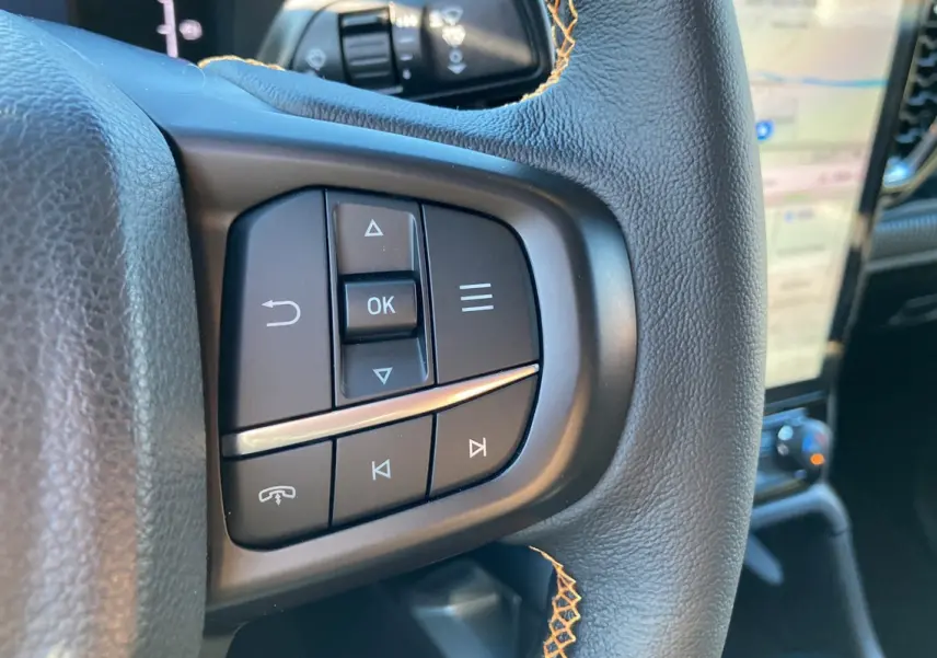 Gros plan sur les commandes gauche du volant cuir noir du Ford Ranger Wildtrak Plus 2025, avec écran tactile en arrière-plan.