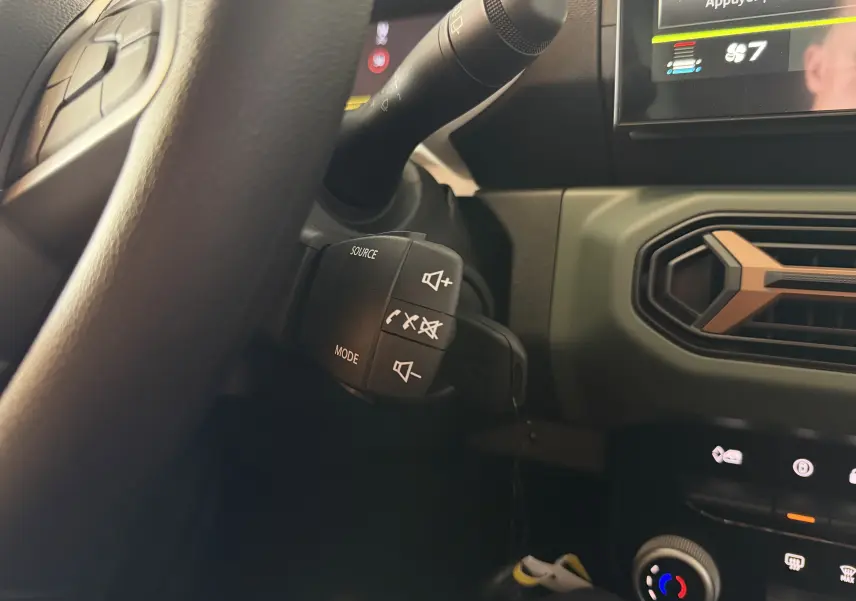 Gros plan sur la commande au volant du Dacia Spring 65 Extreme 2026, avec tableau de bord et aérateur vert olive visibles.