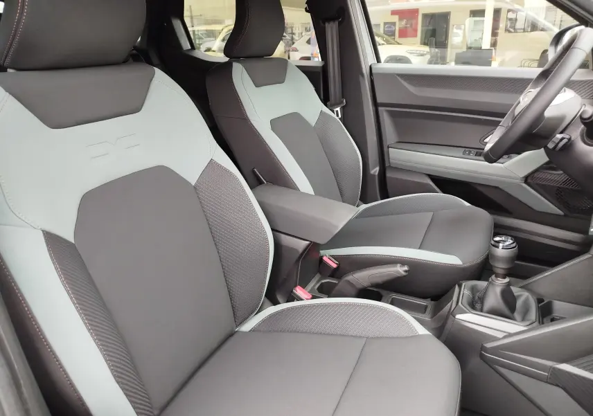 Vue intérieure côté conducteur du Dacia Duster 2025, sièges gris clair et foncé avec surpiqûres orange et levier de vitesse manuel.