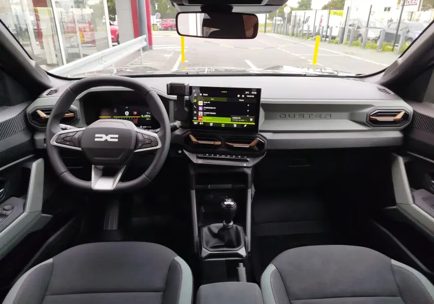 Vue intérieure frontale du tableau de bord du Dacia Duster 2025 avec écran tactile, volant multifonction et levier de vitesse manuel.