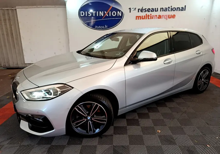 BMW Série 1 gris clair vue 3/4 avant droit, phares LED allumés et jantes sport noires en showroom